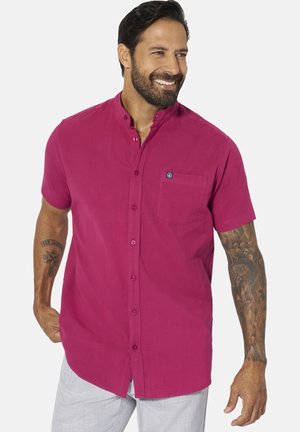 Man smiling, wearing a short-sleeve magenta button-up shirt with mandarin collar and light gray pants, showing tattoos on both arms.