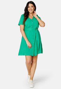 Abito verde a maniche corte con scollatura a V, chiusura a bottoni sul davanti e vita legata. Indossato con sneakers bianche su uno sfondo neutro.