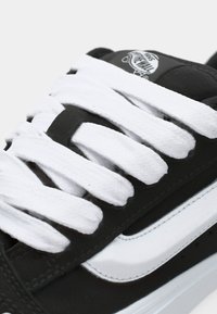 Černé tenisky s bílými tkaničkami, s hladkým svrchním materiálem, charakteristickou nášivkou s logem a kontrastním bílým pruhem na boku.