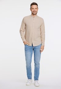 Licht beige overhemd met mandarin kraag, lange mouwen, en een borstzak, gecombineerd met lichtblauwe jeans en witte sneakers.