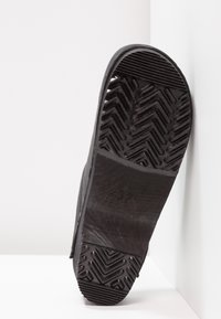 Semelle de chaussure noire avec des motifs de grip en zigzag et linéaires texturés, marquée taille 37, présentée sur un fond de surfaces blanches en angle.
