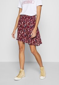 Gonna floreale bordeaux realizzata in tessuto leggero, con bordo arricciato, vita elasticizzata, indossata con una maglietta bianca e sneakers alte beige.