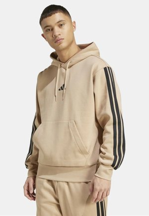 Joven con sudadera con capucha beige y pantalones a juego con rayas negras, de pie contra un fondo claro y liso.