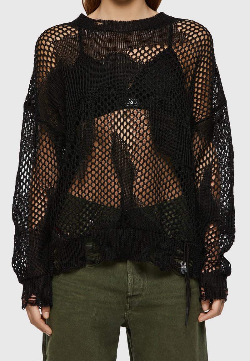 Pull en maille noire présentant une coupe ample avec un large décolleté et des bords effilochés. Le design comporte de larges mailles ouvertes, mettant en valeur les couches sous-jacentes.