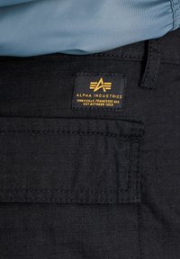 Bolso de pantalón de tejido negro texturizado con parche amarillo de Alpha Industries y parte de tejido azul claro en la parte superior.