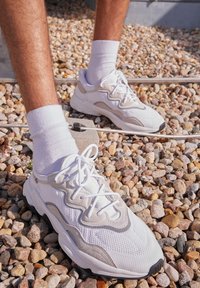 Zapatillas de malla blancas con detalles en gris, suela gruesa y punta redonda. Usadas con calcetines blancos, colocadas sobre una superficie de grava texturizada.