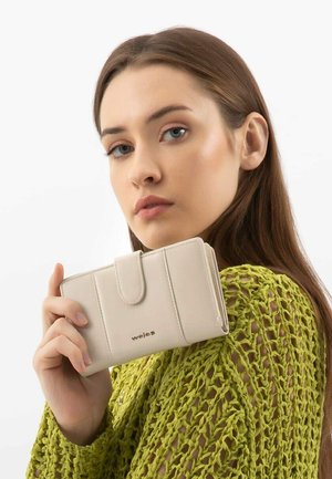 Portafoglio beige chiaro con una texture liscia, caratterizzato da un flap frontale e un branding discreto. Abbinato a un top verde con maglia allentata.