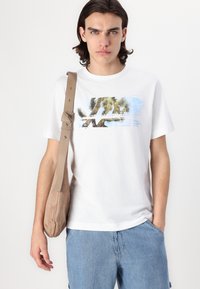 Hvid bomulds T-shirt med et farverigt grafisk design med palmer. Modellen har lyseblå denimshorts og en beige skuldertaske på.