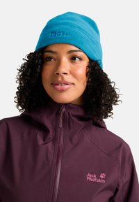 Vrouw met krullend haar, gekleed in een heldere blauwe fleece muts en een paarse Jack Wolfskin zip-up jas, die licht naar de zijkant kijkt.