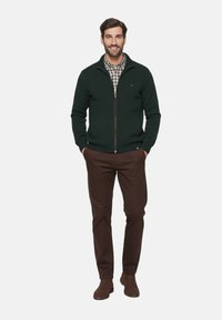 Suéter de cremallera verde oscuro con puños de canalé, llevado sobre una camisa a cuadros, combinado con pantalones marrones y botas marrones. Diseño simple y casual.