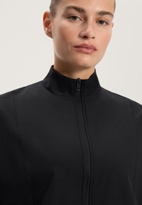 Černá bunda s vysokým límcem vyrobená z hladkého, lehkého materiálu, s předním zipovým zapínáním a minimálním prošíváním.