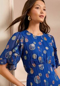 Kraljevo modra obleka z nabranimi kratkimi rokavi in večbarvnimi krožnimi vzorci. Ima oprijet pas in prosojno teksturirano tkanino. Zlati dodatki.