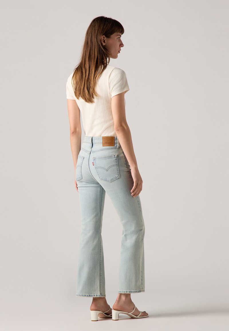 Jean en denim bleu clair avec une coupe slim et des jambes évasées, présentant une pièce en cuir brun à l'arrière de la taille. Porté avec un haut blanc à côtes.