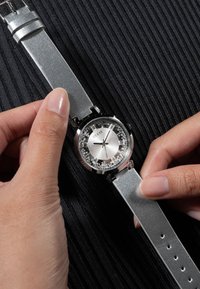 Sølvur med rund urskive, glitrende detaljer omkring skiven og en metallrem, holdt af en hånd mod en struktureret sort baggrund.