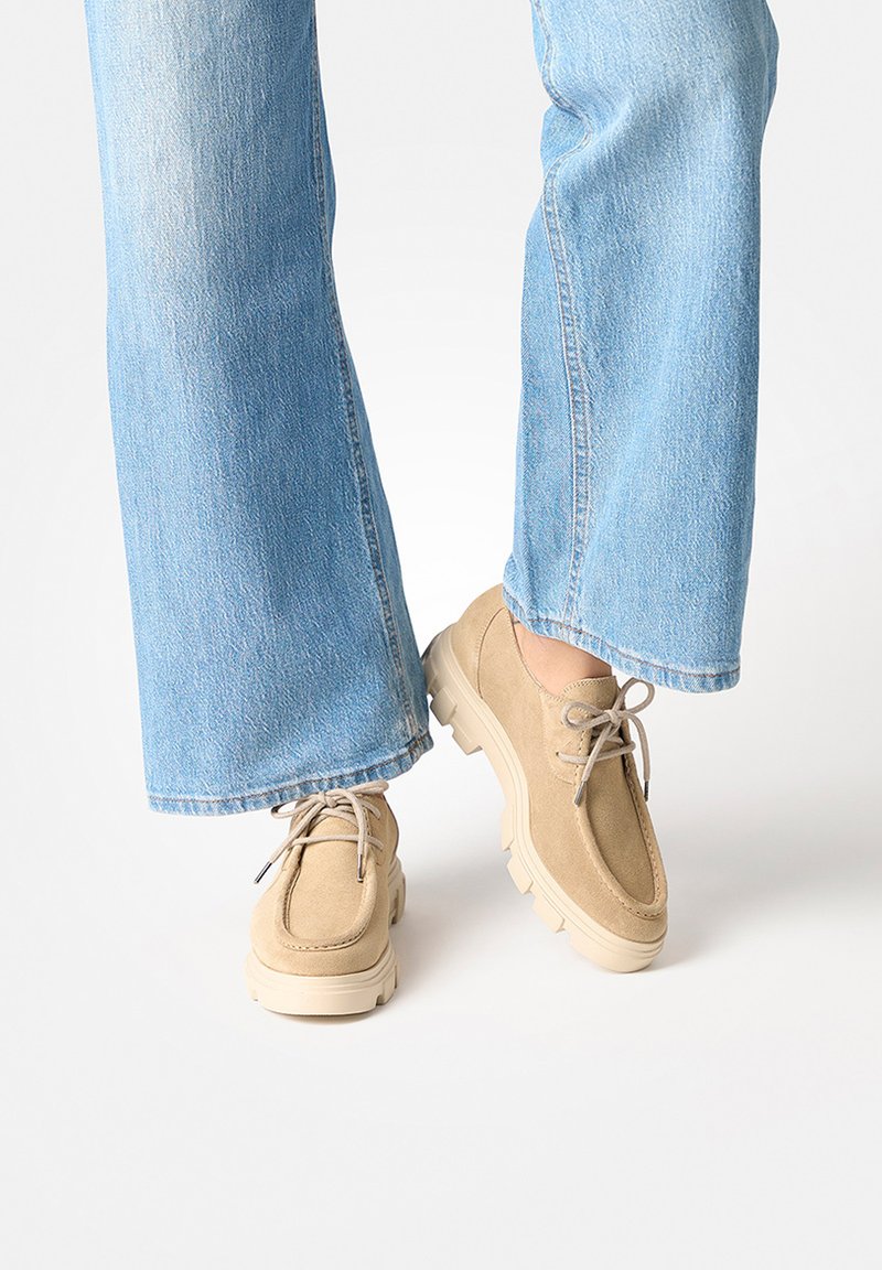 Personne portant un jean bleu clair et des chaussures à lacets en suède beige avec semelles crantées, se tenant sur une surface blanche.