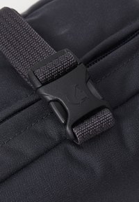 Black fabric bag featuring a textured strap with a plastic buckle, showing fine stitching and a subtle brand logo embossed on the hardware.