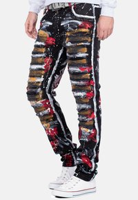 Jean en denim noir avec des zones déchirées, éclaboussures de peinture rouge, dorée et blanche. Design ajusté, bandes latérales et une ceinture texturée.