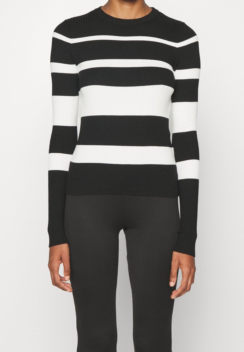 Personne portant un pull à rayures horizontales noires et blanches à manches longues avec un pantalon noir ajusté sur un fond uni.