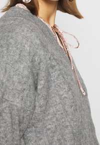 Grauer Strickpullover mit strukturiertem Muster, ausgestattet mit einer rosa Schleife am Hals und einem lockeren Schnitt.
