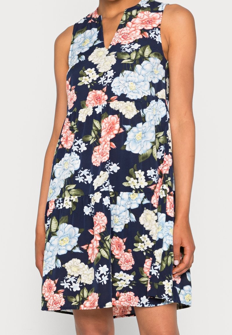 Robe bleu marine sans manches avec col en V, avec un grand imprimé floral rose, blanc, bleu et feuilles vertes, portée par une personne debout.