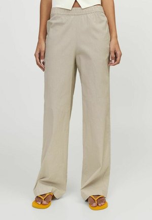 Personne portant un pantalon beige ample avec une taille élastique et des tongs jaunes, debout devant un fond uni.