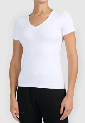 Kobieta ubrana w dopasowaną białą koszulkę z krótkim rękawem i dekoltem w serek oraz czarne spodnie, stojąca na tle jednolitego jasnego tła.