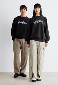 Zwarte sweatshirt met grafische tekst, beige cargobroek met zakken, en witte sneakers, tegen een effen achtergrond.