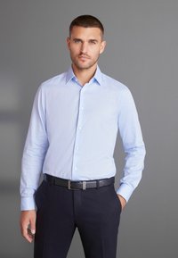 Svijetloplava, košulja dugih rukava s kragnom, dugmadi sprijeda i lagano teksturiranim materijalom, kombinirana s tamnim hlačama i crnim remenom.