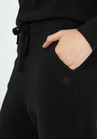 Schwarze Strick-Jogginghosen mit einem Zugbund, seitlichem Taschen und einem dezenten Logo-Patch an der Tasche. Weicher Stoff und taillierter Schnitt.