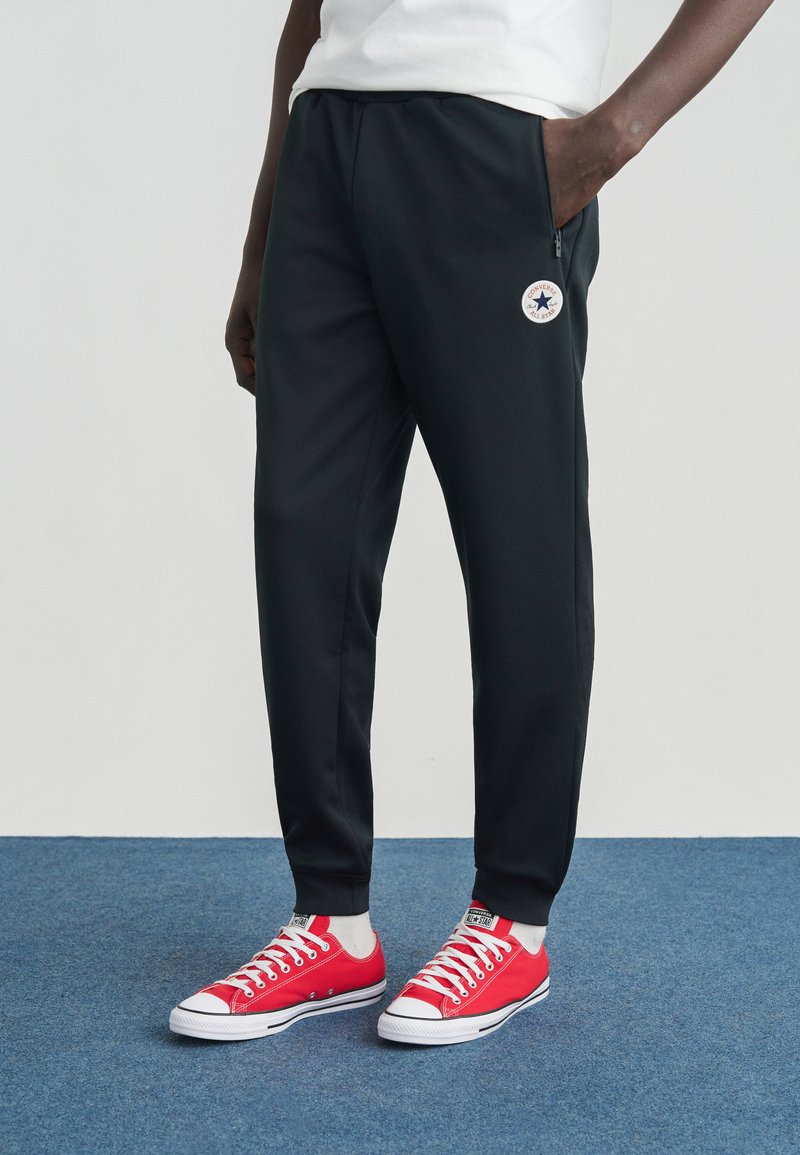 Μαύρο αθλητικό παντελόνι με τσέπη με φερμουάρ και στρογγυλό λογότυπο Converse, σε συνδυασμό με κόκκινα καμβά παπούτσια και ανοιχτόχρωμες κάλτσες αστραγάλου.