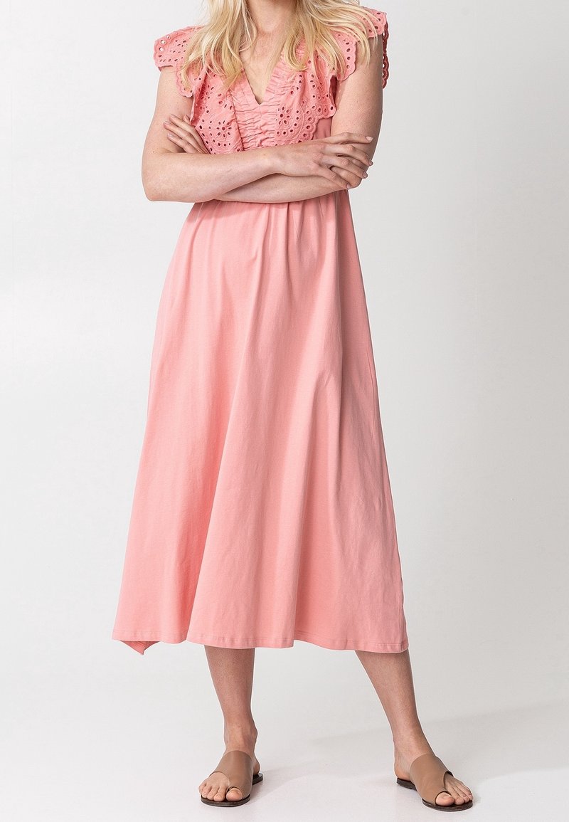 Femme portant une robe midi rose sans manches avec des détails ajourés aux épaules, debout les bras croisés et des sandales plates marron.