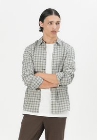 Camisa de cuadros en verde claro, negro y blanco, hecha de una tela suave con botones en la parte delantera, mangas enrolladas, llevada sobre una camiseta blanca básica.