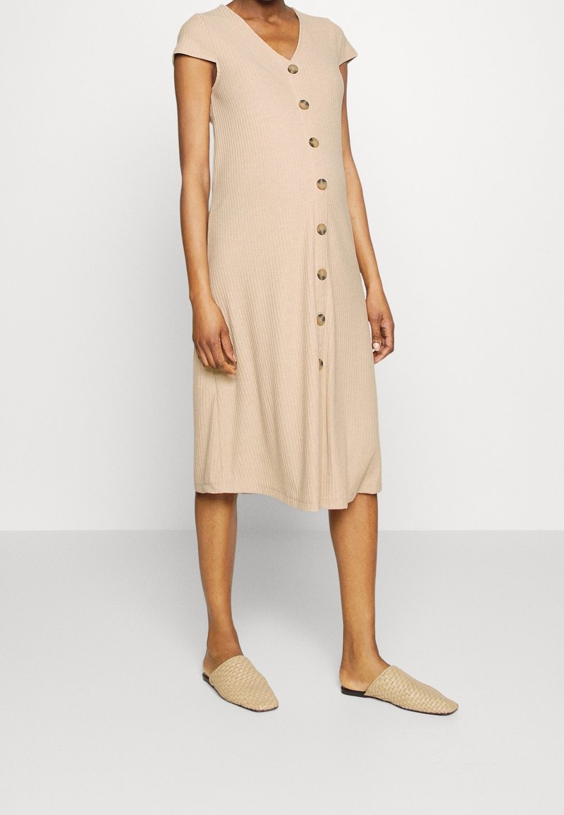 Femme portant une robe midi beige côtelée à manches courtes et à gros boutons sur le devant, associée à des chaussures beiges tissées sans lacets.