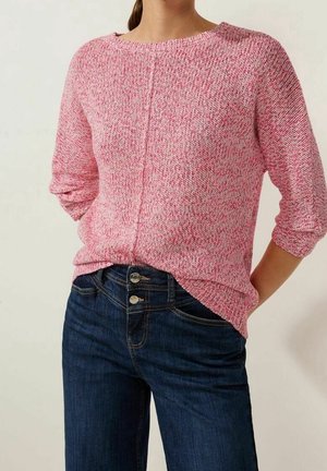 Sweter dzianinowy o teksturowanym wykończeniu w kolorze różowym, z luźnym krojem i okrągłym dekoltem. Nosi się go na ciemnoniebieskie dżinsy z dwoma widocznymi guzikami i klasycznym krojem.