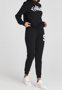 Kobieta ubrana w czarną bluzę z białym nadrukiem oraz czarne spodnie dresowe z białą łatą z numerem "9", w połączeniu z białymi sneakersami na jednolitym tle.