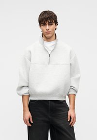 Νέος άνδρας που φοράει ανοιχτό γκρι φούτερ με φερμουάρ μέχρι τη μέση και μαύρα τζιν, με τα μανίκια γυρισμένα, σε απλό ουδέτερο φόντο.