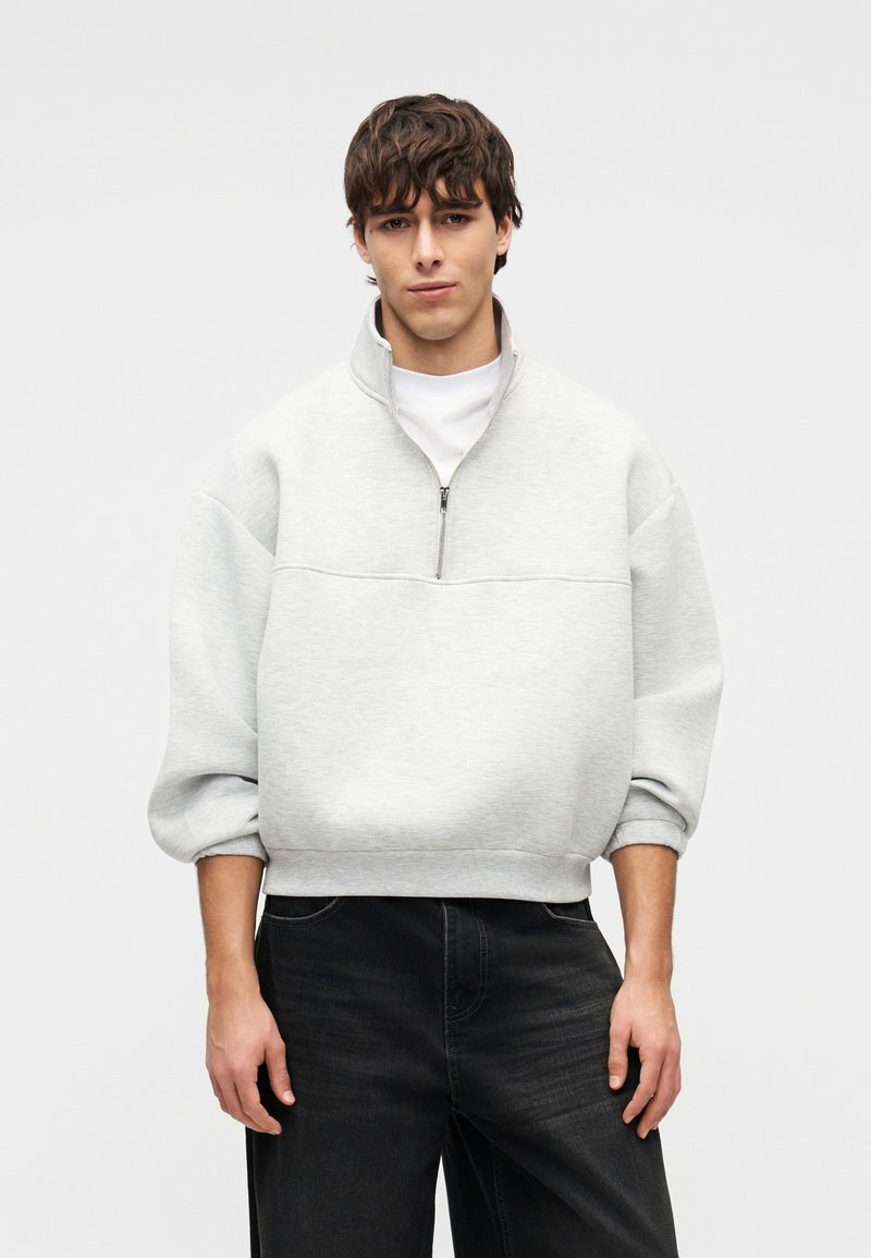 Νέος άνδρας που φοράει ανοιχτό γκρι φούτερ με φερμουάρ μέχρι τη μέση και μαύρα τζιν, με τα μανίκια γυρισμένα, σε απλό ουδέτερο φόντο.