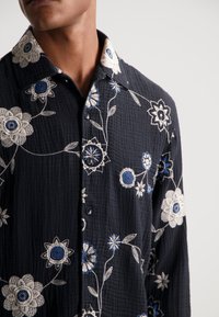 Camisa negra con textura y bordado floral en blanco y azul, que presenta un cuello abotonado y detalles de costuras intrincadas.