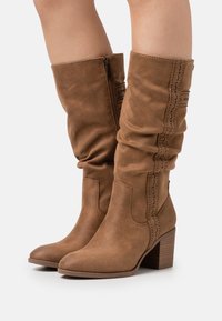 Bottes en daim marron hauteur genou avec un design flou, dotées de coutures décoratives, d'une fermeture éclair sur le côté et d'un talon carré.