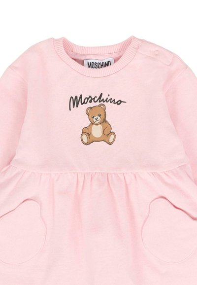 Abito da bambina rosa chiaro con grafica di orsetto, testo "Moschino", tasche a forma di cuore e bottoni a pressione sul colletto della spalla.