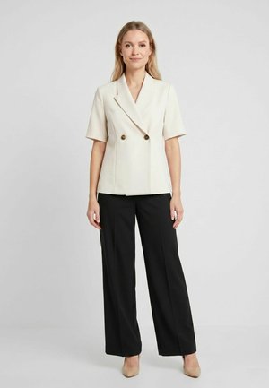 Mujer con blazer crema de manga corta y doble botonadura, pantalones negros de pierna ancha y tacones beige, de pie sobre un fondo sencillo.