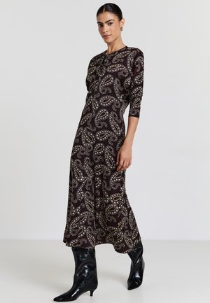 Abito lungo e scuro con un motivo paisley in crema e oro. Presenta maniche a tre quarti e una silhouette fluida. Abbinato a stivali neri fino al ginocchio.