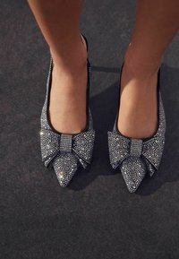 Mustad varvastega lamedad jalanõud, kaunistatud hõbedaste rhinestone'idega ja suure lipsu üksikasjaga ees, millel on sile tekstuur ja moodne disain.