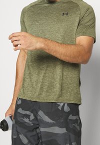 Muži v olivově zeleném tričku Under Armour a černo-šedých maskáčových šortkách, který drží bílou láhev na vodu s černým víčkem.