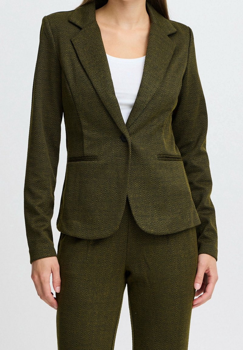 Femme portant un blazer en chevrons vert olive et un pantalon assorti sur un top blanc, debout avec les bras détendus le long du corps.