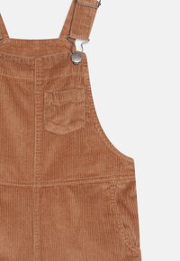 Rudi-brown spalvos švarklų kelnės su reguliuojamais metaliniais sagtimis, viena krūtinės kišenė ir šonkaitymo tekstūra.