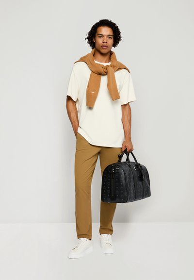 Hombre con camiseta blanca, suéter color camel drapeado sobre los hombros, pantalones color camel, zapatillas blancas, sosteniendo una bolsa negra con estampado frente a un fondo liso.