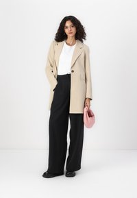 Manteau beige ajusté avec revers crantés, pantalon noir à jambes larges, t-shirt blanc et sac à main rose. Texture lisse avec un design structuré.