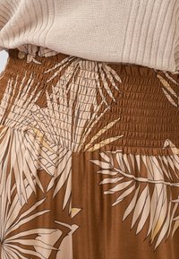 Hnědá sukně se smockovaným pasem a bledým botanickým potiskem. Materiál vypadá lehký a hladký, s volným střihem.