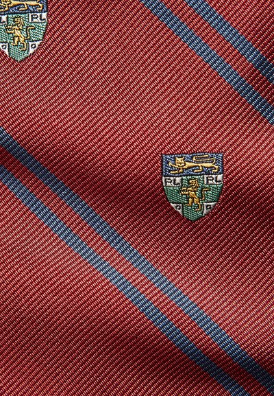 Gravata de seda vermelha com riscas diagonais azuis, apresentando brasões bordados em verde, azul e dourado, exibindo detalhes intrincados e textura.